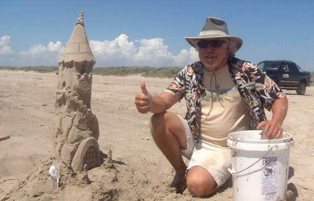 A man in a bucket hat and tropical shirt kneels next to a sandcastle tower and gives a thumbs up. His other hand rests on a white five gallon bucket.