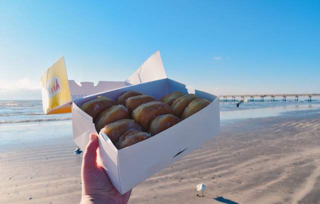 A box of a dozen donuts is held up in front of the beach
