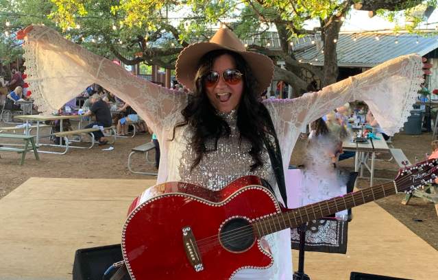 A woman in a white outfit with a red guitar holds her arms out in celebration