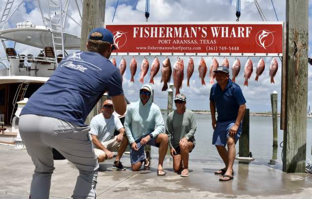 A group of four poses in front of a board of hanging red snapper while a man in blue takes their photo