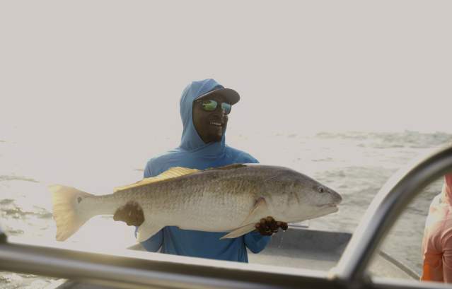 A man in Columbia PFG gear holds up a large black drum fish.