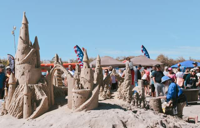 In the foreground, the back of a sandcastle is visible. In the background, the sculptor kneels over his project while a crowd watches and strolls by.