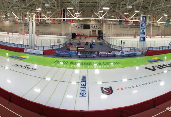 The interior of the Pettit National Ice Center with white ice adorned with decals and lines in the foreground, two ice rinks in the midground and a long tan wall in the background and lights shining down reflecting off the ice throughout.