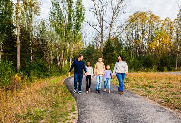 A family of five smiles and walks together along a paved trail at Firefly Grove Park surrounded by colorful autumn trees.
