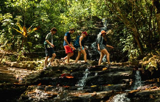 A group of hikers crossing a stream near Pozo Azul Waterfall, surrounded by many trees and nature.