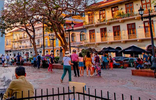 The image shows a vibrant street scene in a historic city. The street is lined with colorful buildings, some with balconies and ornate details. There are people walking around, enjoying the evening. In the background, there is a church with a blue facade and a bell tower.
