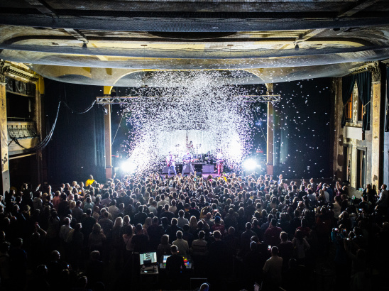 Interior view of Turner Hall ballroom during a live concert, showing a packed crowd of hundreds of people facing an illuminated stage. The historic venue's ornate architecture is visible with decorative columns, balconies, and detailed ceiling work. Dramatic stage lighting creates bright white beams cutting through the dark space, with confetti or snow effects falling from above.