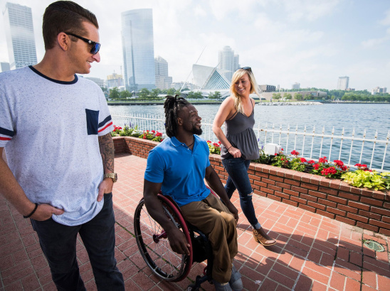 Three friends, including one using a wheelchair, wander along Milwaukee’s lakefront with the skyline in view.