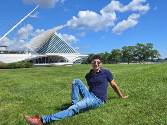 Person sitting relaxed on a grassy lawn in front of the Milwaukee Art Museum on a sunny day with blue skies and scattered clouds.
