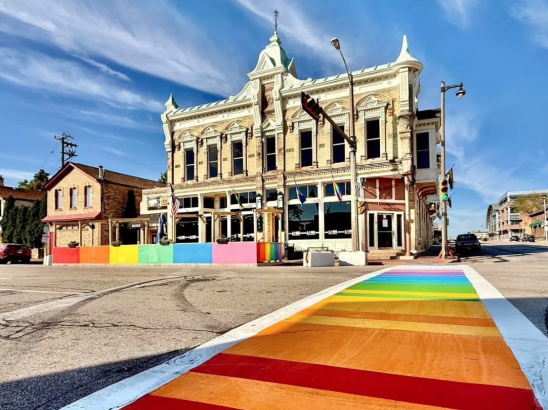 Historic cream-colored brick building with ornate architecture and rainbow pride flags, situated at a street corner with a brightly painted rainbow crosswalk leading toward it under a clear blue sky.