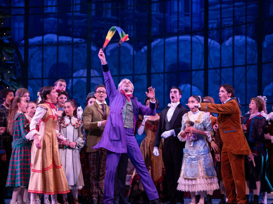 A group of performers dressed in 19th-century costumes gathers on stage during a festive scene. At the center, a man in a bright purple suit joyfully raises a colorful toy or ribbon stick above his head, surrounded by children and adults in elegant period attire. The background features tall windows and a large Christmas tree, evoking a holiday celebration.