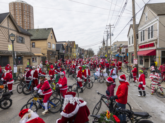 A large group of cyclists dressed in Santa Claus outfits fills a Milwaukee street during a festive holiday bike ride, surrounded by local shops and winter scenery.