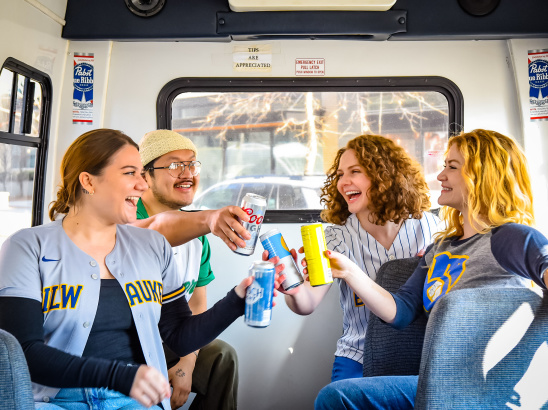 Four friends sitting inside a shuttle bus, smiling and clinking their drinks together. Two are wearing Milwaukee sports jerseys, creating a festive and celebratory atmosphere.