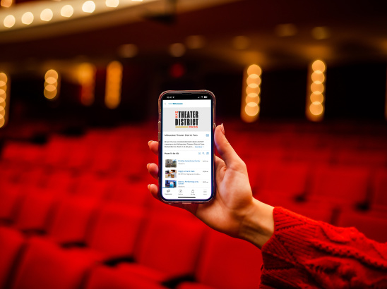 Hand holding up cellphone displaying homescreen of Milwaukee Theater District Pass in front of blurry theater background