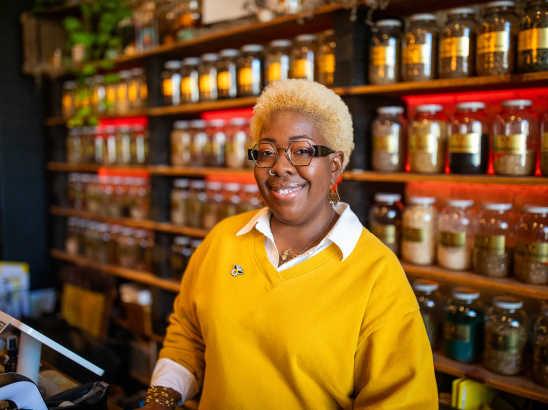 Angela Mallett is smiling, standing behind the counter of HoneyBee Sage. She's wearing a bright yellow sweater over a white collared shirt, along with gold jewelry including earrings and a necklace. Behind her are wooden shelves lined with numerous glass jars containing various spices, herbs, or other ingredients with labels.