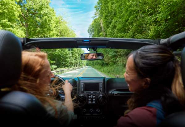 Two women in a corvette driving down an open road with green trees framing the road