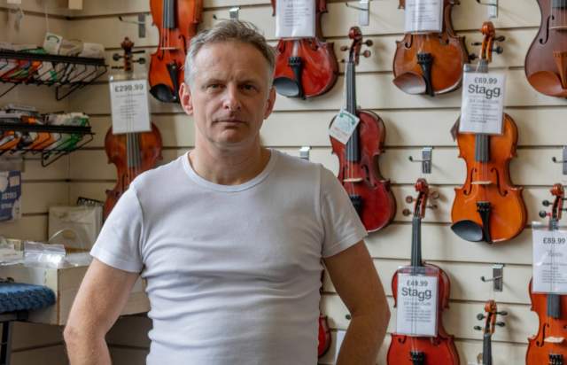 John Roadhouse Jr pictured in front of a wall of violins in his music shop