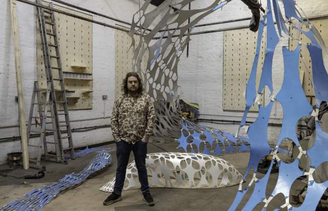 Liam, an artist and sculptor stood behind a blud sculpture