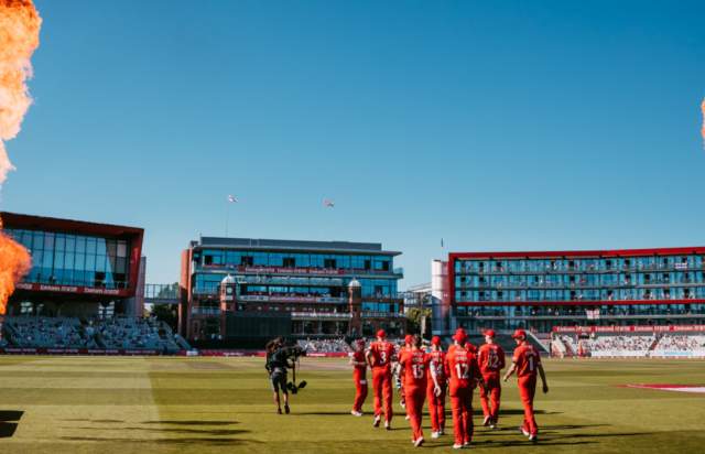Cricket team walking on to pitch with fire on a sunny day