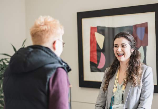 A young white woman with long wavy brown hair smiling in front of an abstract art picture whilst chatting to another white woman who has her back to the camera