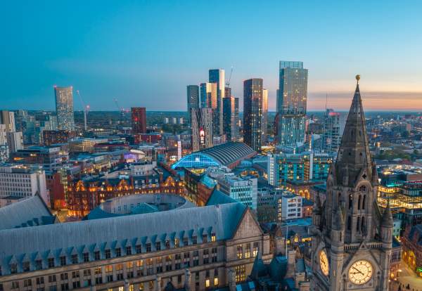A panoramic evening view of Manchester city centre at dusk, with the illuminated clock tower of Manchester Town Hall Extension in the foreground and Manchester Central Convention Complex at the heart of the skyline. Modern high-rise towers at Deansgate Square and surrounding city lights glow against a blue twilight sky, showcasing the contrast between Manchester's historic and contemporary architecture.