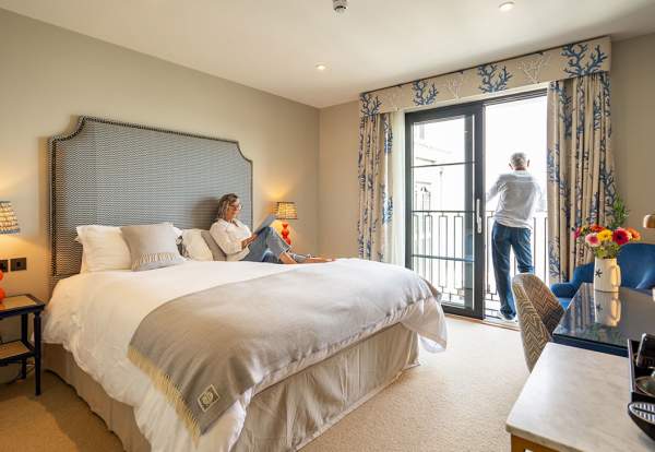A man and a woman in a hotel room. Woman reading on the bed, man looking out of the window.