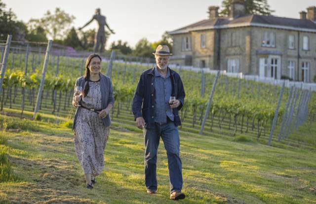 A couple walk through the vines, with Leonardslee House and a sculpture in the background