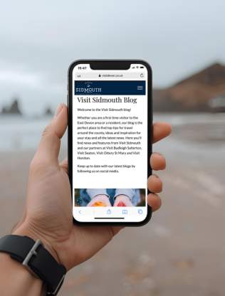 A man is holiding his phone while on the beach in Sidmouth. He is reading the Visit Sidmouth Blog