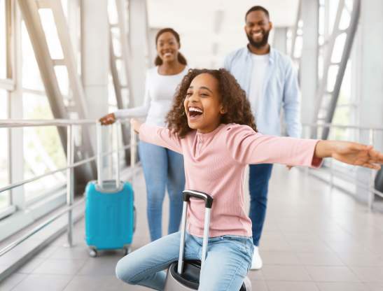 Summer Vacation Concept. Portrait of cheerful African American girl having fun, sitting on luggage and spreading hands, pretending plane flying. Family of three people going on journey