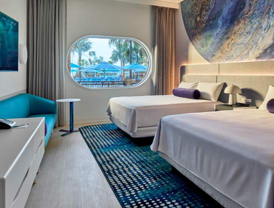 A double queen room at Universal Stella Nova Resort, featuring two queen beds, a dresser, couch, TV, galaxy-inspired wall mural and and a spaceship-style window.