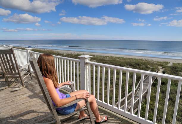 Person sitting on the upper deck of an oceanfront vacation rental in the Brunswick Islands, overlooking sandy dunes and gentle ocean waves under a partly cloudy sky.