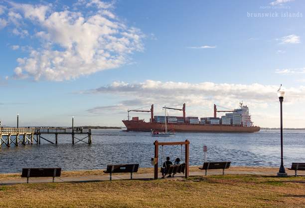 a couple sitting on the swinging benches at Southport Waterfront Park and a shipping vessel and a sailboat go by