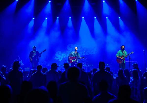 Band playing on a stage with shadows of fans in the foreground. The whole image is lit by blue lights from above.