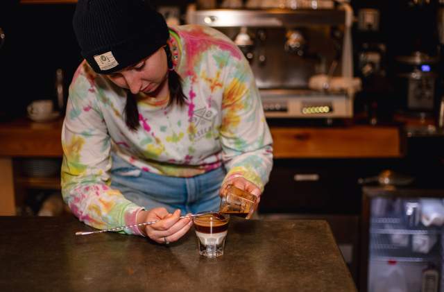 A barista in a tie-dye long sleeve and black beanie on her head, pours espresso into a glass.