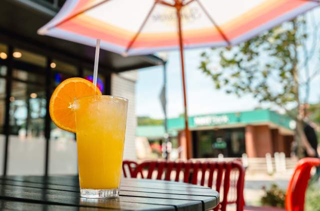 A refreshing orange cocktail garnished with a slice of orange, served in a tall glass with a straw on an outdoor patio table under a colorful umbrella, with a vibrant downtown setting in the background.
