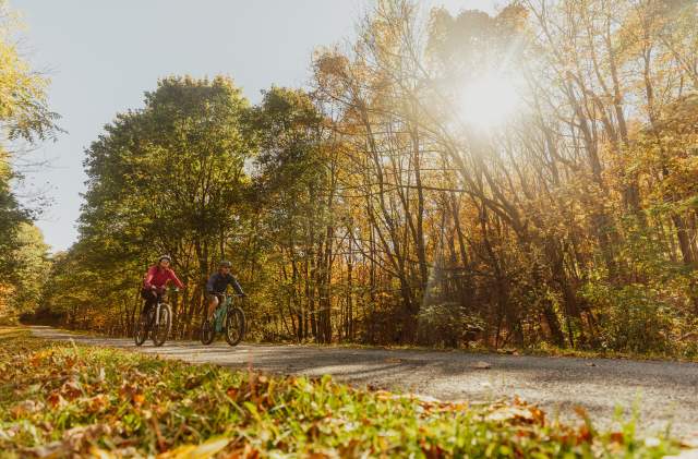 A man and a woman are each on their own bike. peddling along the Great Allegheny Passage in the fall.
