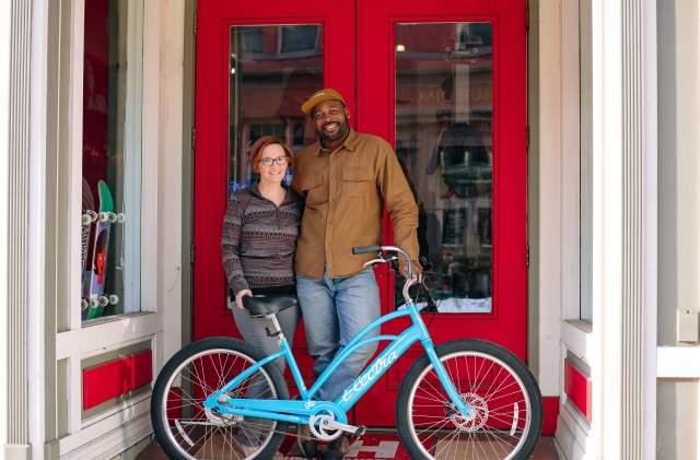 A man and woman stand in front of a red door holding a bright blue bike.