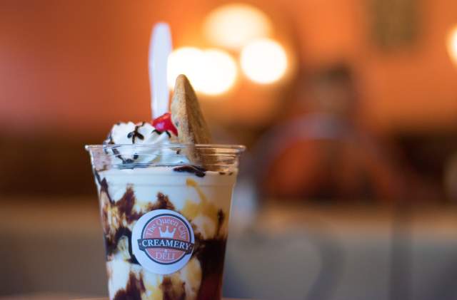 A cup of Queen City Creamery vanilla ice cream with hot fudge and topped with whip cream, a cheery, and a large chocolate chip cookie.