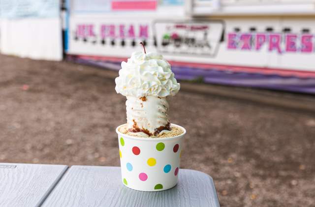 A green, pink, yellow, and blue polka dot cup with vanilla ice cream, whipped cream, and a cherry sits on top of table.