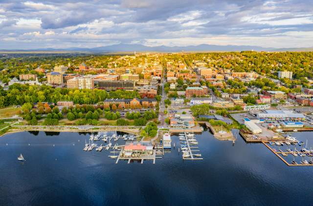 Aerial view of Burlington by a lakeshore with marinas, docks, and numerous buildings. Greenery surrounds the area, and mountains are visible in the background.