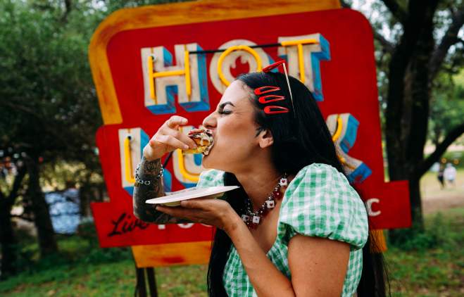 Image of a girl taking a big bite of food at Hot Luck Festival with the red Hot Luck sign behind her.