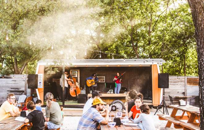 People sitting on wooden picnic benches under shaded oak trees at Sour Duck, listening to a live music set.