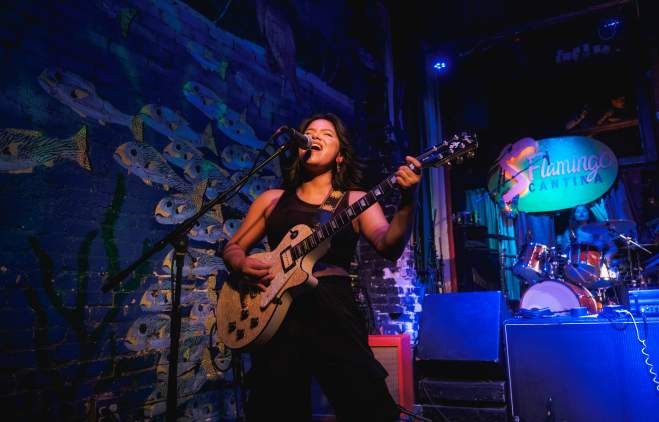Woman standing on a stage painted with an irredescent fish backdrop, playing the electric guitar and singing into a microphone.