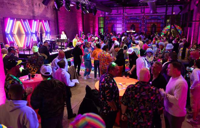 Group of people in a venue with a neon backdrop on a stage, enjoying a conference after party.