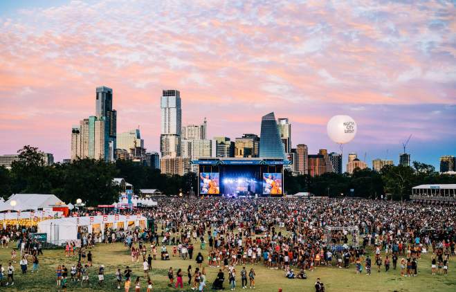 Aerial shot of Zilker Park during ACL festival with a sea of fans crowding the main stage, a cotton candy colored sky with the Austin skyline as the backdrop.