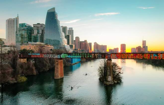 Kayakers paddling under a bridge with grafitti on Lady Bird Lake while the sun sets against Downtown Austin buildings.