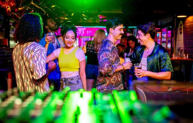 Couples dancing with cocktails in hand in front of a DJ booth with neon lights in an intimate space.