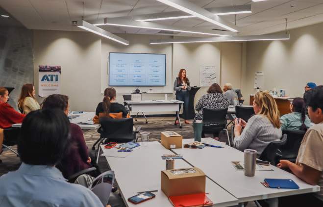 People gathered in a classroom setting, looking at a teacher at the front of the room.