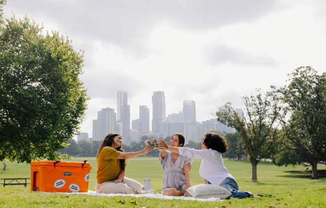 Three women sitting on a picnic blanket cheersing with drinks in Zilker Park.