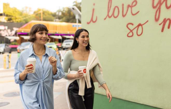 Two women walking past "I love you so much." mural with to-go coffee cups in hand.